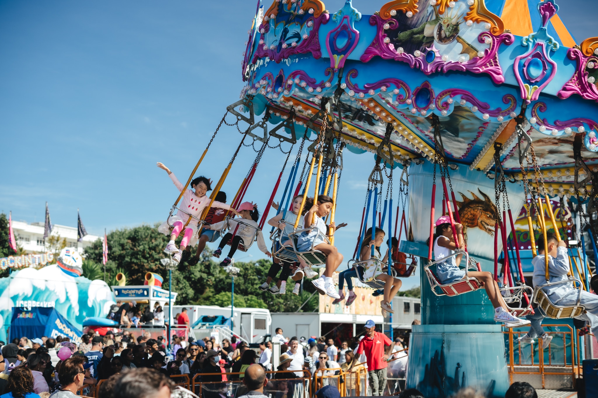 Children laughing and swinging through the air on a colourful carousel swing ride at a sunny outdoor fair, blue sky overhead.